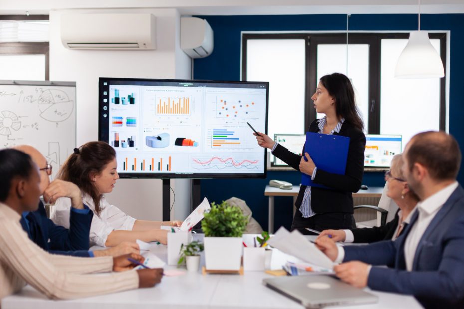 Жінка в офісі із синьою папкою в руках, що демонструє колегам презентацію: на екрані діаграми та графіки різних видів та кольорів. За столом сидять двоє чоловіків та троє жінок, частина з них – в окулярах, дехто літнього віку, хтось читає інформацію з листка, хтось дивиться в екран